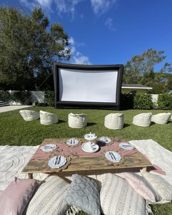 Immerse yourself in the ultimate Movie Night Extravaganza, where a sprawling outdoor cinema awaits! Picture a massive screen set against the backdrop of vibrant green grass, enhanced by plush bean bags thoughtfully arranged for premium comfort and unparalleled viewing pleasure. In the foreground, a charming low wooden table is elegantly adorned with place settings and cozy cushions, creating an inviting atmosphere. Surrounded by lush greenery and basking beneath a pristine blue sky, this idyllic setup promises an unforgettable cinematic experience under the twinkling stars—perfect for movie lovers seeking magic in the great outdoors.