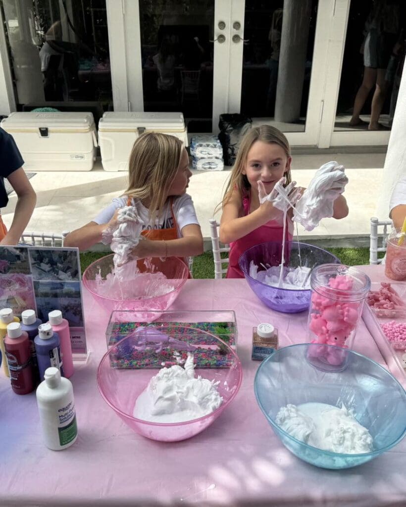 In a vibrant scene of creativity and fun, two excited children engage in playful exploration with rainbow-colored slime at a bustling craft table. Surrounded by an assortment of bowls brimming with crafting materials and various bottles filled with glittery supplies, their laughter echoes the enthusiasm of a Movie Night Extravaganza. This imaginative activity sparks joy and inspires endless possibilities for kids' DIY projects.