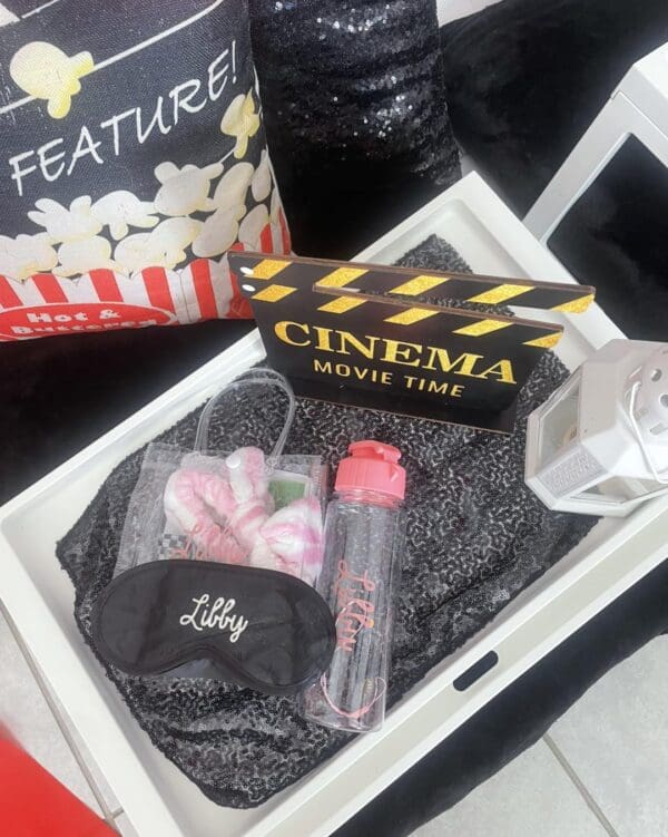 Immerse yourself in the ultimate Movie Night Extravaganza with this chic and stylish setup. A sleek tray, adorned with a classic clapperboard, invites cinematic flair, while a cozy eye mask promises relaxation. Quench your thirst from an elegantly designed water bottle as you cuddle up with an adorable plush toy companion. Organize your movie essentials in charming baskets placed on a soft gray blanket that enhances the scene's cozy ambiance. Add to the cinematic charm with a delightfully themed popcorn pillow and enchanting decorative lights nearby, creating the perfect atmosphere for a memorable night of films and fun. Unwind and enjoy your dream movie marathon in style!