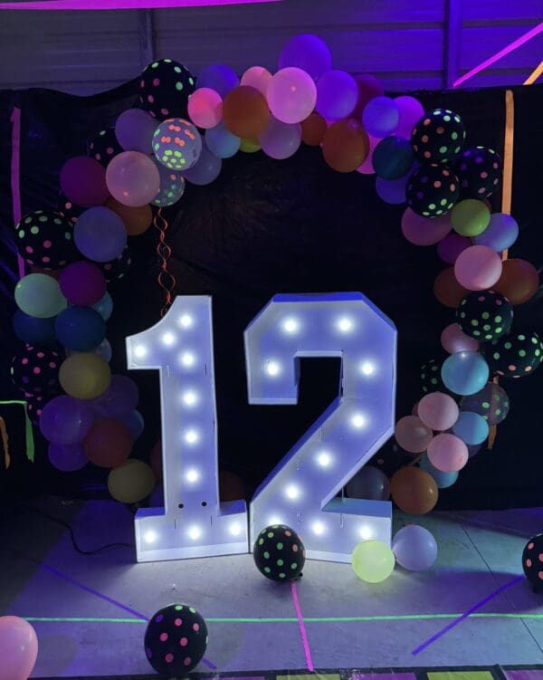 A vibrant "12" sign aglow with dazzling light takes center stage, elegantly framed by a stunning arch of multicolored balloons, creating the perfect festive atmosphere for an unforgettable Movie Night Extravaganza. Nestled in this dimly lit venue, the display captivates guests with a sense of cinematic wonder and celebration. Keywords: Movie Night Extravaganza, illuminated 12 sign, colorful balloon arch, festive atmosphere.
