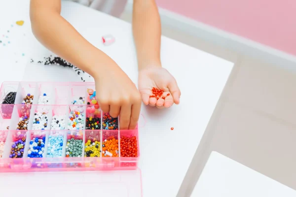 A child's hands selecting red beads from a colorful assortment for a Jewelry Art Party project in a storage box on a white table.