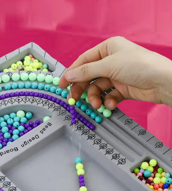 A person's hand threading colorful beads onto a string using a Jewelry Art Party with various compartments filled with beads to create unique jewelry.