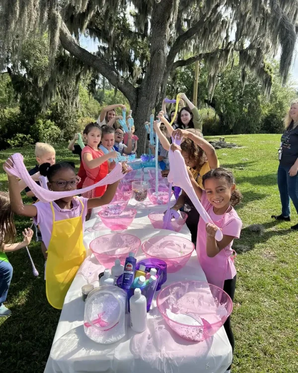 Children are gathered outside at a long table for a Slime Party, mixing various colorful ingredients in bowls. Two adults accompany them, while a large tree provides shade in the background.