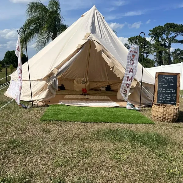 A large, white canvas tent is set up outdoors on a grassy area. Inside, the tent is furnished with cushions and a rug, perfect for a Harry Potter Themed Party. A chalkboard sign and decorative banner featuring Hogwarts motifs are placed nearby.