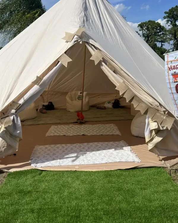 A white tent with an open entrance is set up on a grass lawn. Inside, white cushioned seats and throw blankets are arranged, perfect for a Harry Potter Themed Party. The tent is decorated with small banners.