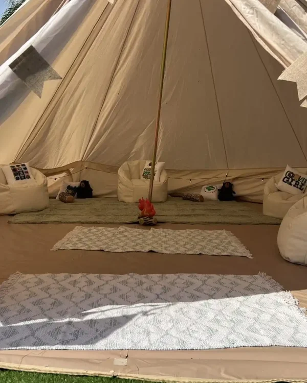 A cozy glamping tent with white beanbag chairs arranged around a small central fire pit, various cushions, and rugs on the ground. The tent is well-lit, creating a warm and inviting atmosphere perfect for a Harry Potter Themed Party.