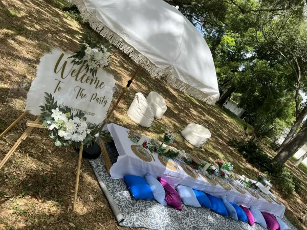 Outdoor picnic setup for a Harry Potter Themed Party with a decorated sign welcoming guests, a table set with plates and floral arrangements, blue and pink cushions on a patterned rug, and a large white umbrella for shade.