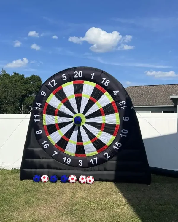An inflatable dartboard with numbered sections is set up outdoors on the grass, with six colorful soccer balls ready for fun. Perfect for a Harry Potter Themed Party, a white fence and house provide the backdrop under a clear blue sky.