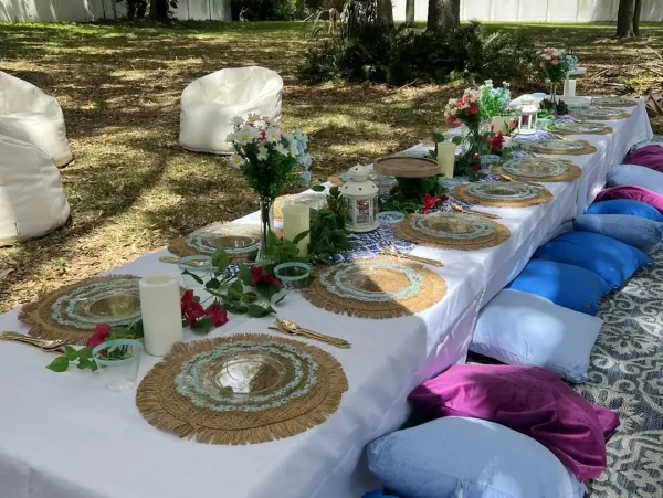 A long, low table is set outdoors with decorative plates, floral centerpieces, candles, and surrounded by colorful cushions and bean bags for seating. The setting, perfect for a Harry Potter Themed Party, is in a shaded, grassy area.