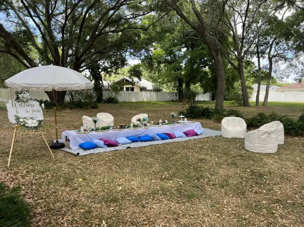 A picnic setup in a backyard with a low table, cushions, and two bean bag chairs. A sign on an easel next to a white umbrella reads "Welcome to the Harry Potter Themed Party." Trees and houses are in the background, setting the perfect scene for a Harry Potter themed party.