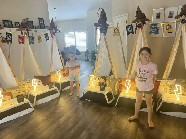 Two children stand beside indoor tents adorned for a Harry Potter Themed Party, complete with house banners and sorting hats. Each tent has a glowing lightning bolt light in front.