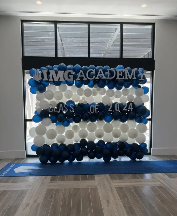 A balloon display featuring blue and white balloons forms a backdrop with the text, "IMG Academy" and "Class of 2024" inside a modern room with natural light from windows above. The scene is reminiscent of a Harry Potter Themed Party, blending magic with celebration.
