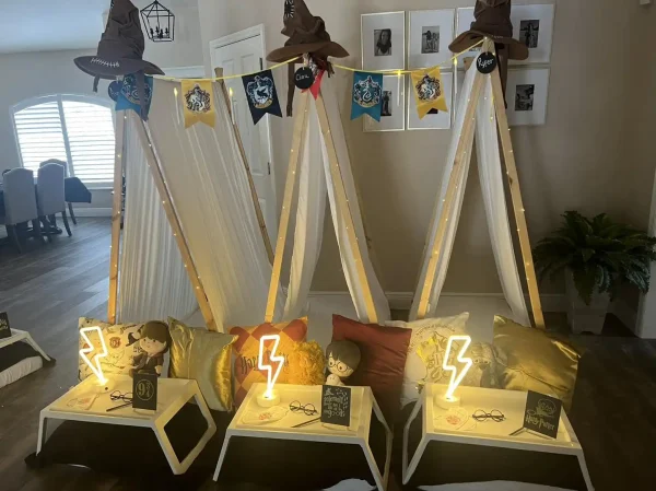 Three indoor teepees with Harry Potter-themed decorations, including pillows and banners, and illuminated lightning bolt signs. Small tables in front hold themed books and glasses, creating the perfect setting for a Harry Potter Themed Party.