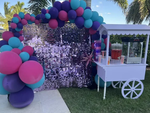 A purple and pink balloon arch birthday party rental that surrounds a shimmering sequin backdrop next to a white cart with a popcorn machine for a slumber party
