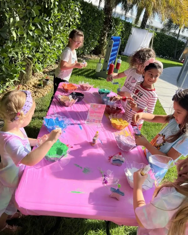 Children gathered around a pink table outdoors, engaging in arts and crafts with various colorful materials and supplies, all centered around a Taylor Swift Theme.