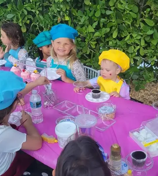 Children wearing colorful hats sit around a decorated table with a pink tablecloth, cupcakes, and crafting supplies at an outdoor Taylor Swift Theme-themed party.