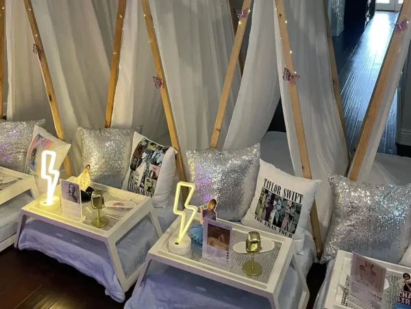 Indoor setup with Taylor Swift Theme decorated with sheer white fabric and fairy lights. Each teepee has a small table with sequin pillows, magazines, and lightning-shaped lamps, creating a cozy lounge area.