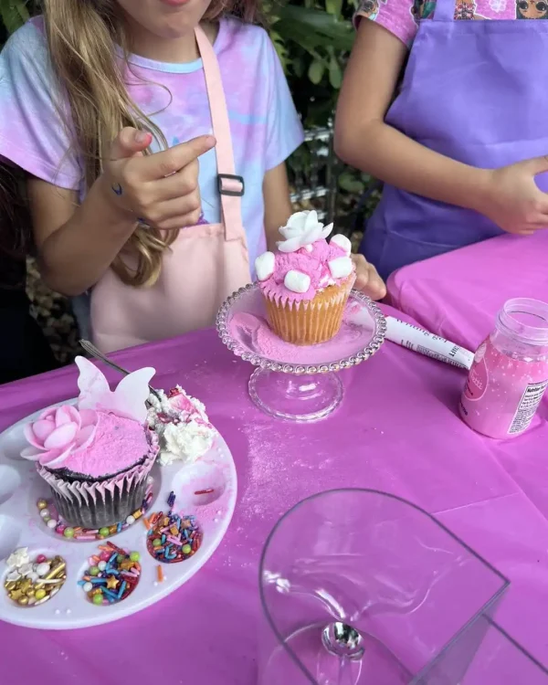 Two children at a table with pink cupcakes decorated with white and pink frosting, sprinkles, and marshmallows. One child points at a cupcake on a stand. The Taylor Swift Theme is evident amid the baking supplies scattered on the table.