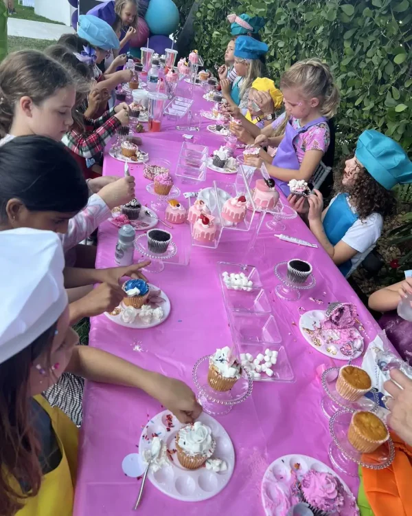 Children gathered around a long table covered with a pink tablecloth, decorating cupcakes with different toppings such as whipped cream and sprinkles. Some children are wearing chef hats and aprons, perfectly complementing the Taylor Swift Theme of the party.