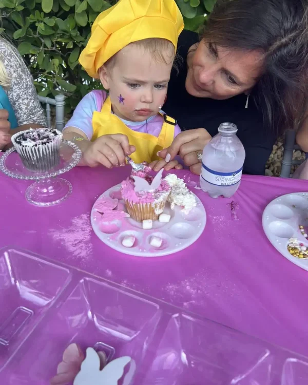 A child, wearing a yellow hat and purple shirt, decorates a cupcake at a Taylor Swift Theme-themed table covered with a pink tablecloth. An adult watches and assists. Various cupcake toppings and a water bottle are on the table.