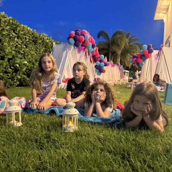 A group of young girls sits on a blanket on the lawn in the evening. Small lanterns twinkle in front while tents with balloon decorations, set up for a *Taylor Swift Theme*, form a magical backdrop.