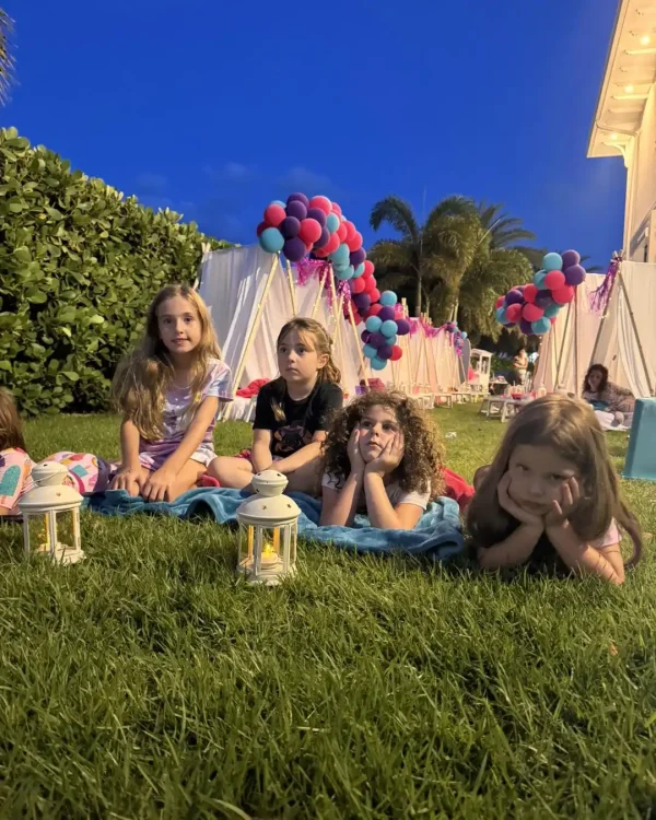 A group of young girls sits on a blanket on the lawn in the evening. Small lanterns twinkle in front while tents with balloon decorations, set up for a *Taylor Swift Theme*, form a magical backdrop.