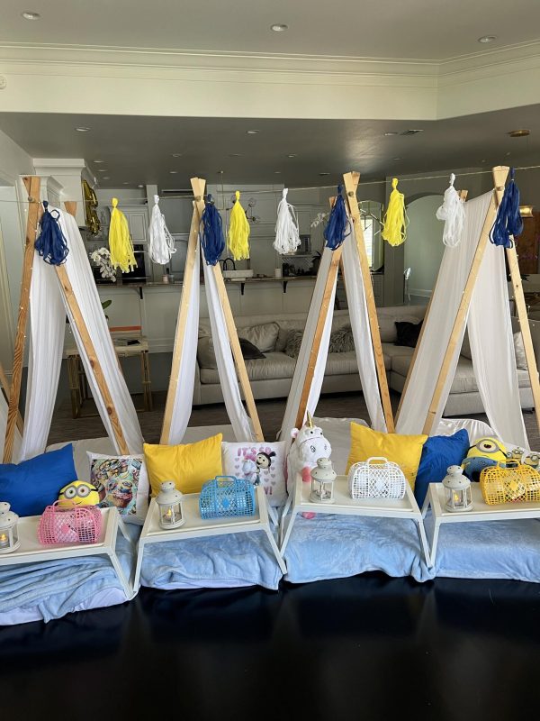 Four indoor teepees arranged with colorful pillows and lanterns are set up in a living room, adorned with yellow, blue, and white decorative tassels hanging above them. The cozy setting feels like it was designed on auto-draft but brings an effortless charm to the space.