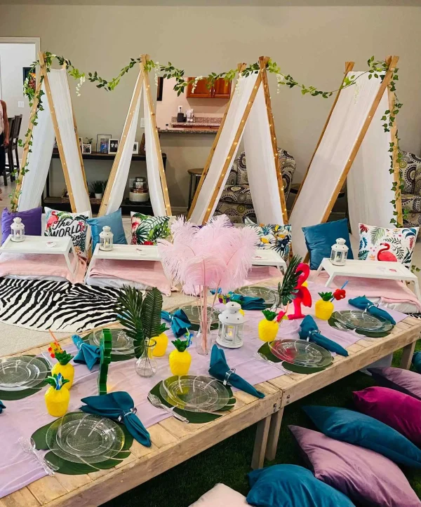 A brightly decorated indoor teepee setup with vibrant pillows, colorful flamingo and pineapple-themed plates, teal napkins, and feathers in the centerpiece. The teepees are adorned with hanging foliage, creating a playful and inviting home ambiance.