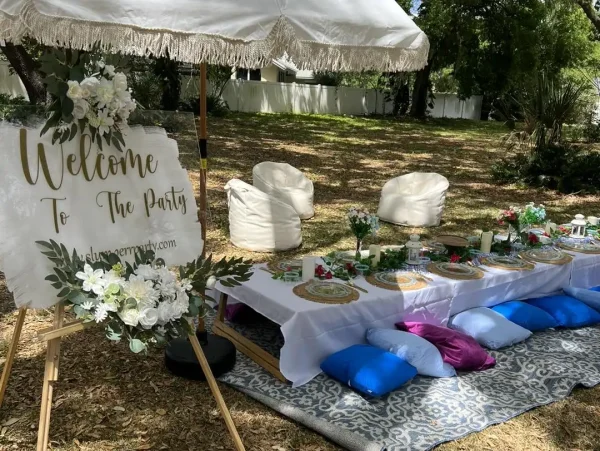 A picnic setup with a white-themed table under a large umbrella. The table is surrounded by blue and white cushions and adorned with floral arrangements, creating a cozy home-like atmosphere. A sign reads, "Welcome to the Party.