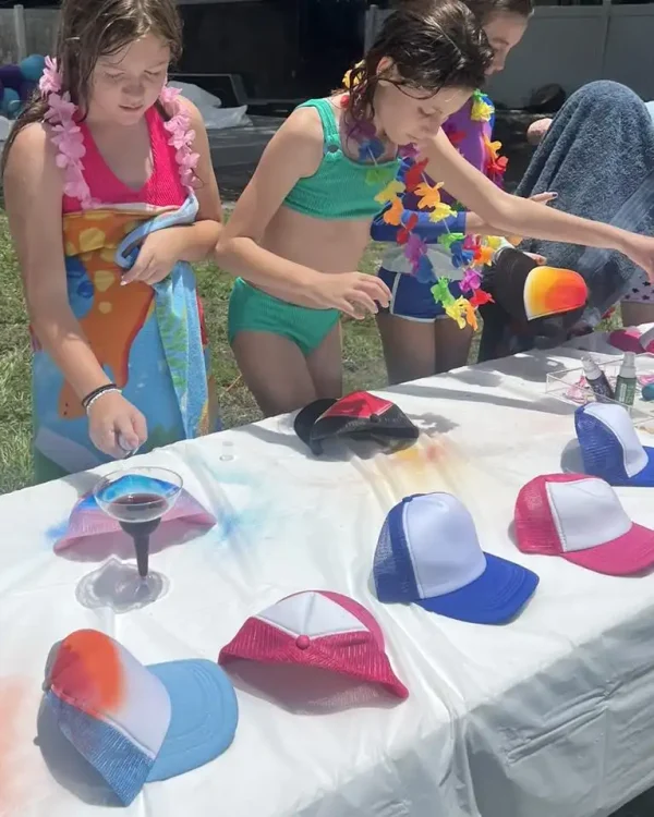 Two children are decorating baseball caps at a table outdoors, reminiscent of a Luxury Soft Play adventure. The table, draped in white cloth, showcases an array of colorful caps. One child wields a spray bottle while the other carefully adjusts a cap, making each creation unique and luxurious.