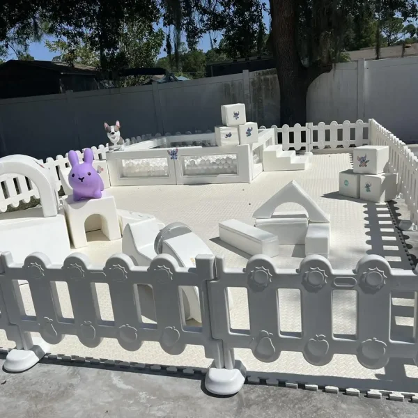 A fenced play area featuring white geometric climbing structures and a variety of stuffed animals, including a purple bunny, is set up outdoors under trees. The Luxury Soft Play Packages ensure both fun and safety for your little ones.