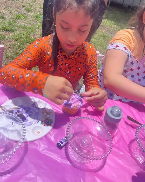 A child, dressed in an orange long-sleeve shirt, decorates a cupcake at a table outdoors. The table is adorned with a pink tablecloth and items from the Luxury Soft Play Packages, exuding creativity. Another person sits beside them.