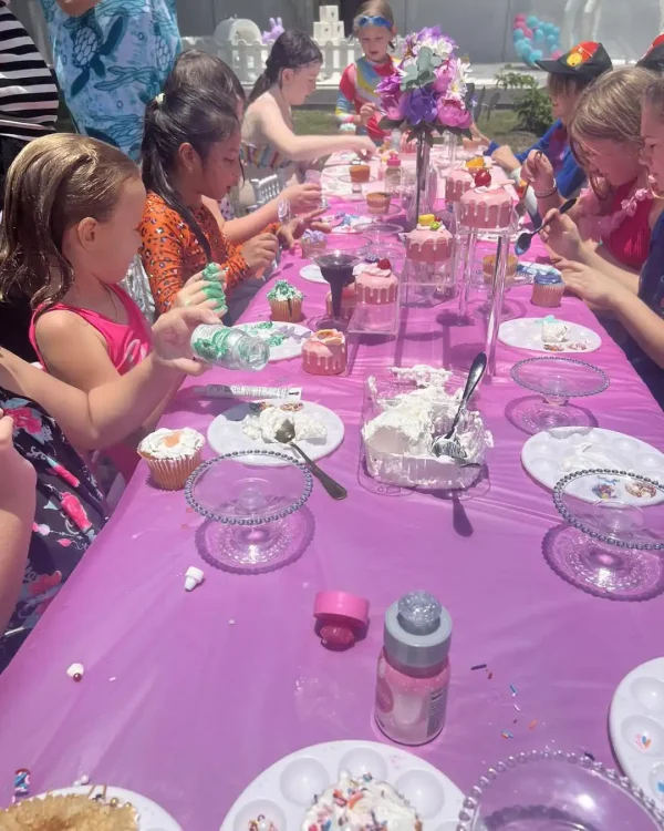 A group of children gathered around a long table adorned with a luxurious purple tablecloth from the Luxury Soft Play Packages, decorating cupcakes with various toppings at an outdoor party.