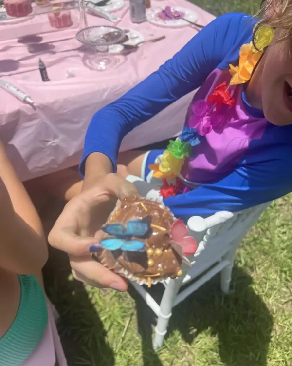 A child holding a decorated cupcake with a blue butterfly candy stands in front of a table adorned with a pink tablecloth and luxurious assorted dessert items. In the background, other children can be seen enjoying the Luxury Soft Play Packages.