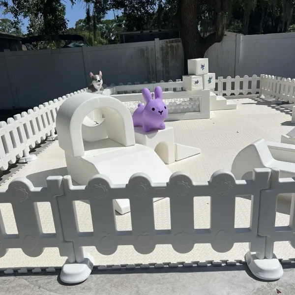 The Luxury Soft Play Package features a fenced-in soft play area with white plastic barriers containing various white play structures and a large purple bunny toy. A small dog-like figure stands on one of the structures, while trees in the background add a blend of luxury and tranquility, perfect for any child's adventure.