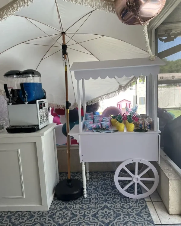 A luxury soft play package features a white drink and candy cart adorned with pineapple cups, set under a large umbrella on a stylish patterned floor. A nearby slushie machine offers blue and white beverages, enhancing the whimsical atmosphere.
