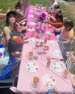 Children seated around a long, outdoor table adorned with a pink tablecloth and flowers are joyfully participating in a luxurious cupcake decorating activity. This enchanting scene is part of the Luxury Soft Play Packages, which also include soft play areas for endless fun.