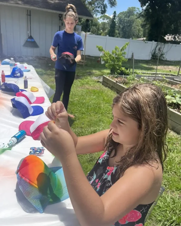 Two people enjoy decorating trucker hats outdoors with products from the Luxury Soft Play Packages. In the foreground, a girl delicately holds a thread, while an adult in the background neatly arranges hats and paints on a table. Garden beds and a fence are visible, crafting an inviting setting that perfectly blends creativity with the luxury of nature.