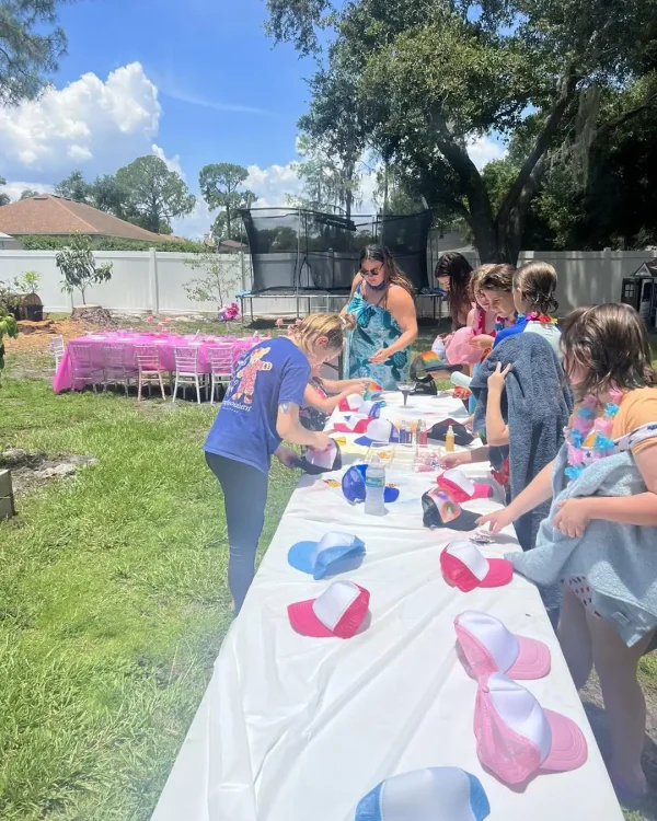 Children are decorating hats at a long table outdoors on a sunny day, supervised by an adult. In the background, more tables with pink covers are visible, hinting at Luxury Soft Play Packages. Trees and houses complete the charming scene.