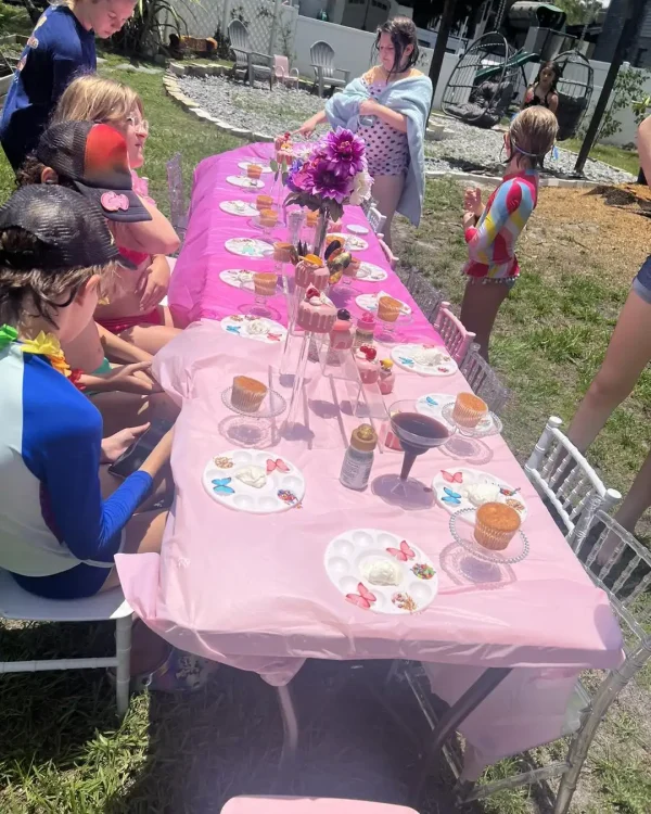 Children in swimwear gather around a long table adorned with pink tablecloths, flowers, and crafting supplies, enjoying a touch of luxury at an outdoor gathering featuring the Luxury Soft Play Packages.