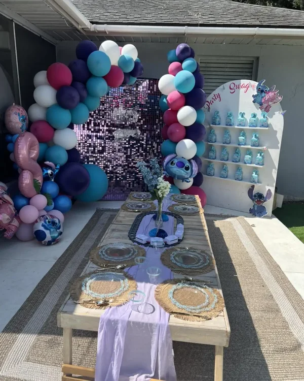 A kids' party setup features a table with purple decor and stitched place settings, a balloon arch with pink, blue, and purple balloons, and a backdrop adorned with luxury soft play packages displaying party favors on shelves decorated with Stitch.