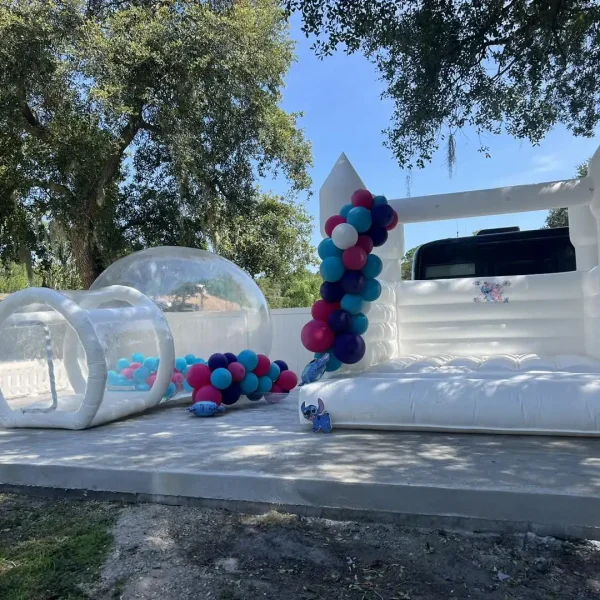 An outdoor setup at home features a white inflatable bounce house and a transparent inflatable tunnel, accentuated by balloon decorations in pink, blue, and white, all under the serene canopy of trees.