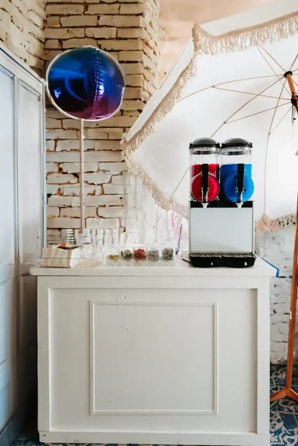 A white counter featuring a drink dispenser filled with red and blue beverages, a stack of cups, and a large decorative balloon set against a white brick wall—perfect for capturing stunning shots with our Event Photography Packages.