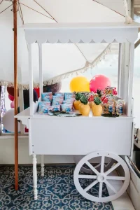 A white cart under an umbrella, adorned with various snacks, drinks, and pineapples, and accompanied by colorful balloons, creating a perfect setting for our Event Photography Packages.