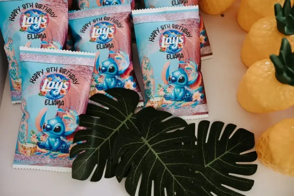 Bags of Lay's chips featuring a Stitch-themed design from the movie "Lilo & Stitch" with "Happy 8th Birthday Eliana" printed on them are beautifully arranged on a table adorned with artificial tropical leaves, captured perfectly through our Event Photography Packages.