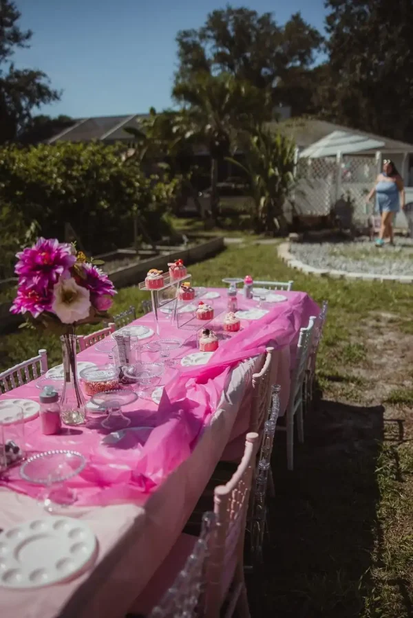 A long table set for an outdoor party is beautifully adorned with a pink tablecloth, flowers, and cupcakes. In the background, a person stands near a garden area and a white gazebo—perfect for capturing memorable moments with our **Event Photography Packages**.