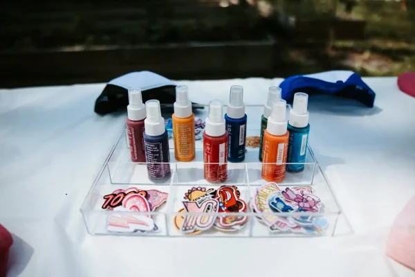 A clear organizer tray from our Event Photography Packages holds six vibrant spray paint bottles and an assortment of colorful patches on a table, ideal for enhancing your event photography setups.