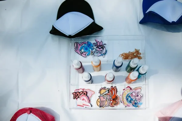 Various colorful patches, bottles of paint, and a black-and-white cap are displayed on a white table, perfect for capturing the essence of Event Photography Packages.