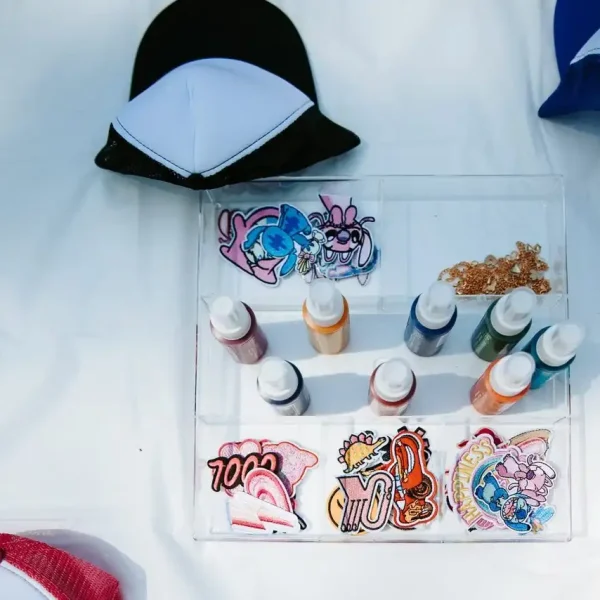 Various colorful patches, bottles of paint, and a black-and-white cap are displayed on a white table, perfect for capturing the essence of Event Photography Packages.