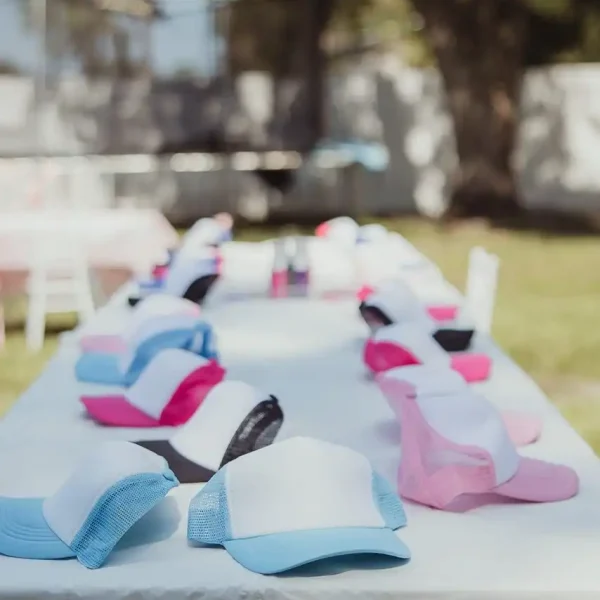 A long table outdoors showcases a vibrant array of trucker hats in blue, pink, white, and black while trees bask in the sunlight in the background. Capture every cherished moment of this colorful scene with our Event Photography Packages.